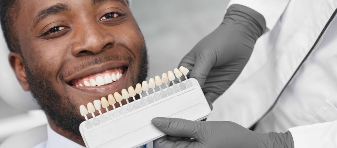 Young african man looking at camera and smiling while female dentist keeping range of fillings in clinic. Happy patient enjoying white beautiful smile while visiting dentist office. Concept of cure.